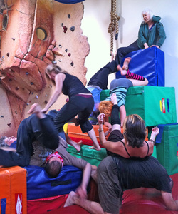 Six dancers on a jumbled tower od gymnastic blocks. On person balanced on another's back, one is slithering down from the top, where another is perched.
