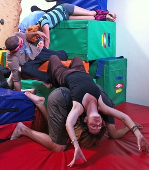 Photo of 4 people dancing on a jumbled tower of large colorful gymnastic blocks. In the forground, one person is supported by another who is on all fours. Behind, a blindfolded dancer reaches up toward s a person slithering down from the top.